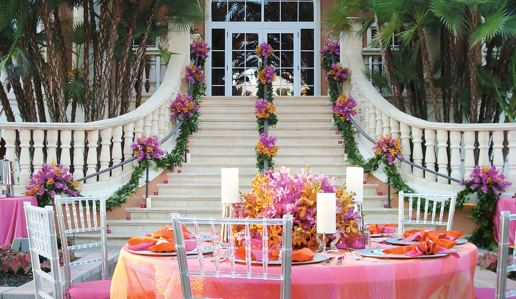West Bay Club's Special Events Elegant outdoor staircase decorated with colorful flowers behind a bright event table with candles and acrylic chairs.